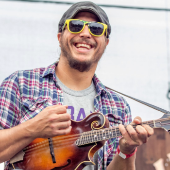 A smiling man in sunglasses and a plaid shirt plays a mandolin outdoors.