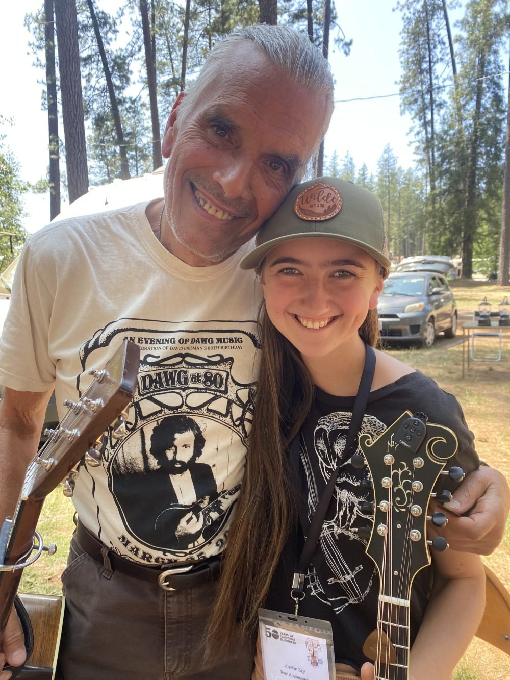 Two people stand outdoors, smiling at the camera. Both hold stringed instruments, and there are trees, cars, and camping chairs in the background.