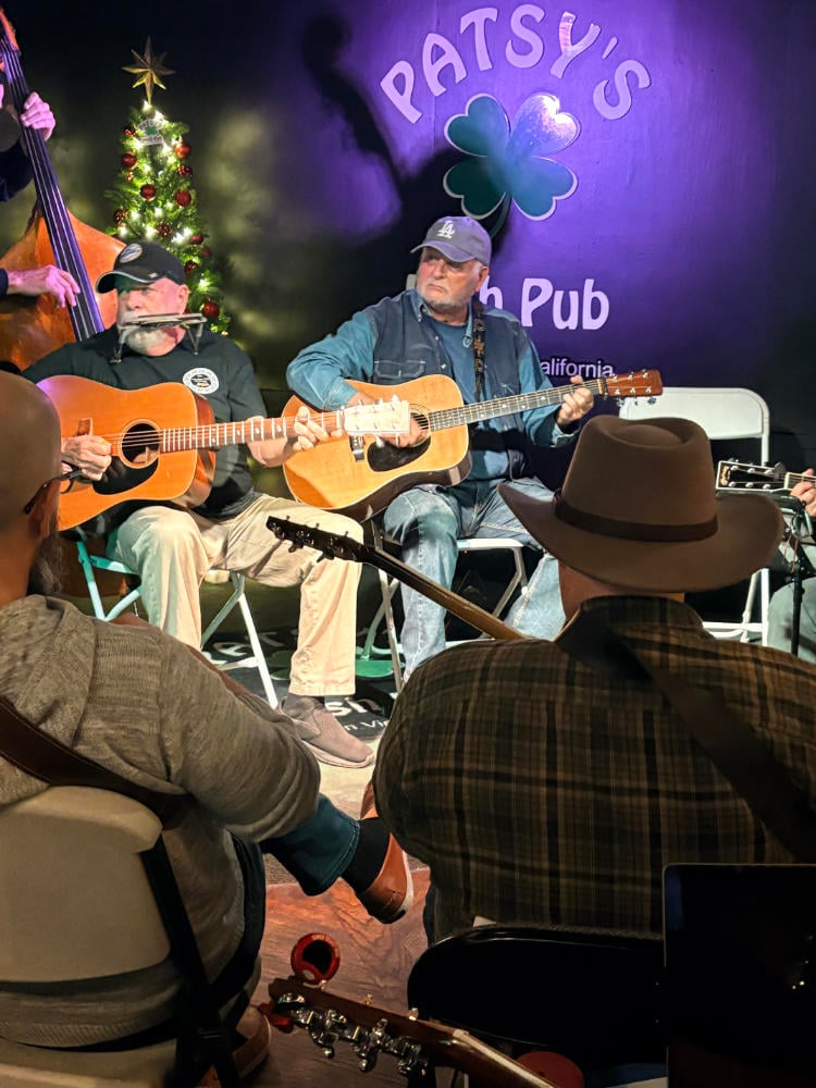 A group of musicians play acoustic guitars on stage at Patsy's Irish Pub, with a lit Christmas tree in the background.