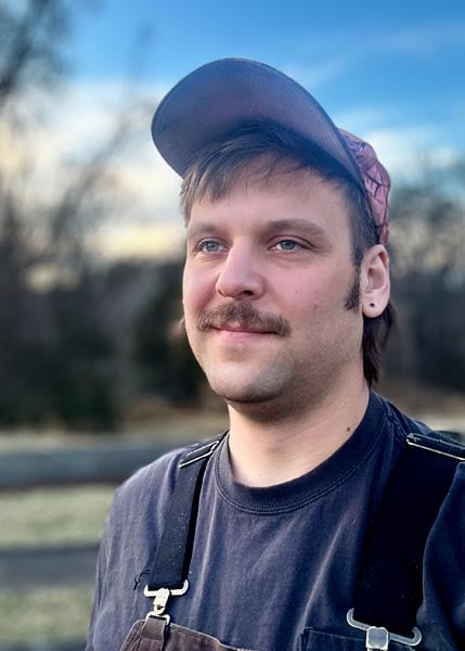 A person with a mustache wearing a dark shirt, overalls, and a cap stands outdoors with trees and a blue sky in the background.