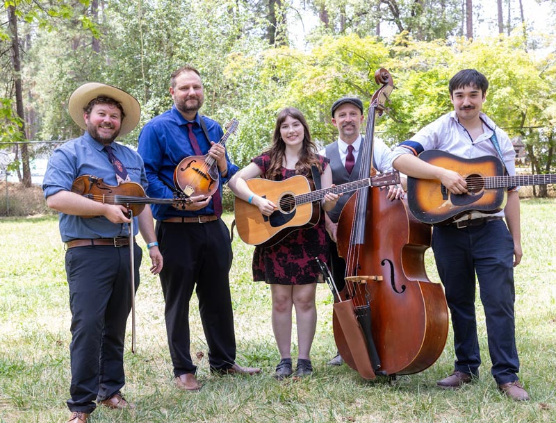 Bay-Area-Special_opt Five people stand outdoors with musical instruments including guitars, a mandolin, and an upright bass, posing for a group photo.
