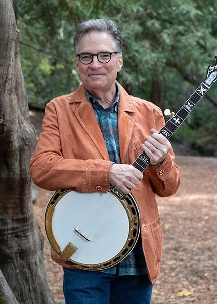 Bill-Evans-SMC2026 A man wearing glasses and an orange jacket stands outdoors holding a banjo, with trees and a dirt path in the background.