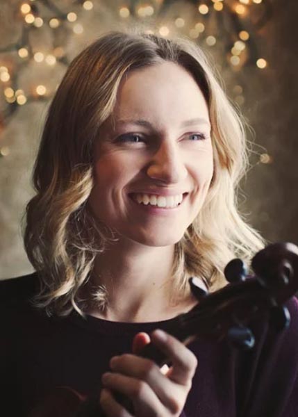 A woman with shoulder-length blonde hair smiles while holding a violin, with string lights softly glowing in the background.