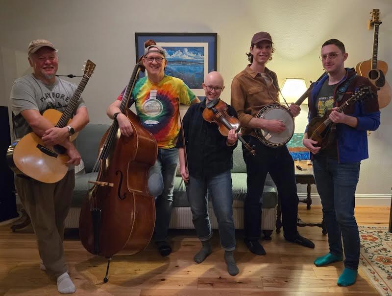 Critical-Grass_OPT Five people stand indoors with musical instruments including guitar, upright bass, violin, banjo, and mandolin. A guitar hangs on the wall behind them and a painting is visible.