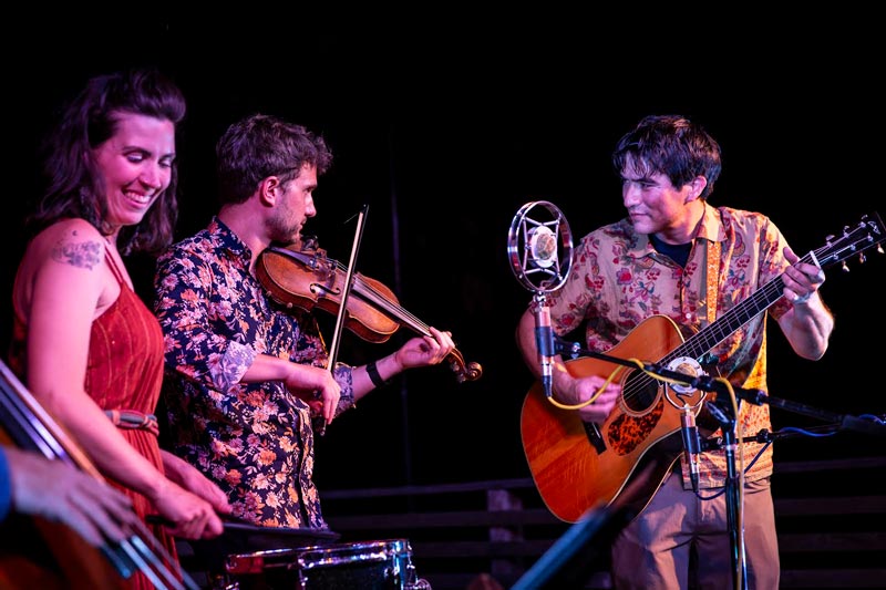 Deep-Thicket-Dwellers_opt Three musicians perform on stage, playing a violin and guitar, with a microphone in front; one is smiling. They wear patterned shirts and are illuminated by stage lighting.