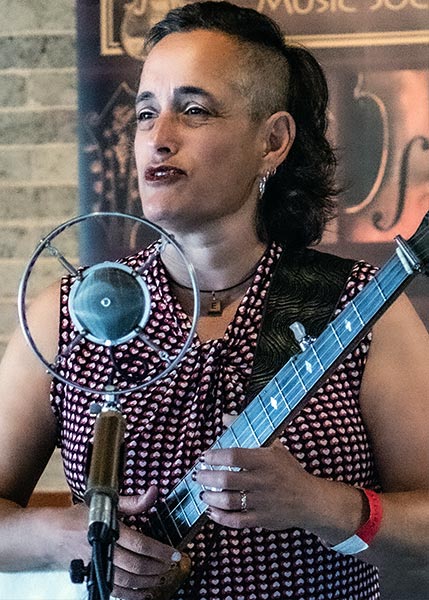 Evie-Ladin-SMC2026 Person with short, curly hair and a patterned sleeveless shirt stands at a microphone, holding a banjo, with a blurred banner and brick wall in the background.