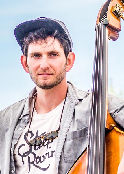 Forrest-Marowitz-SMC2026 A man wearing a cap and sunglasses around his neck stands outdoors, holding an upright bass.