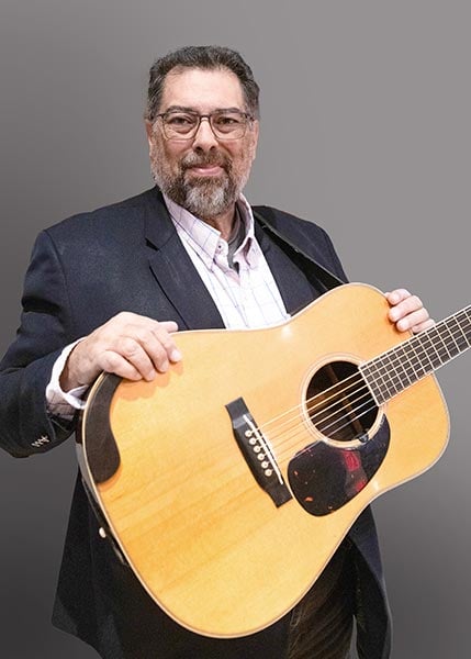 A man with glasses and a beard stands holding an acoustic guitar, wearing a dark blazer over a light shirt, against a plain gray background.