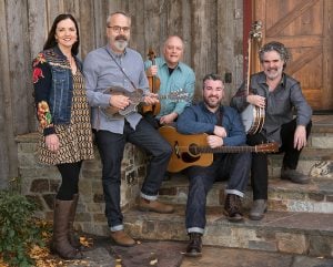 Five adults pose on outdoor steps with musical instruments, including a banjo, guitar, violin, and mandolin, in front of a wooden building.