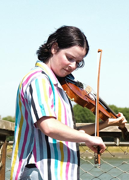 Tatiana-Hargreaves-SMC2026 Person with short dark hair plays a violin outdoors, wearing a colorful striped shirt, with a blurred background of trees and sky.