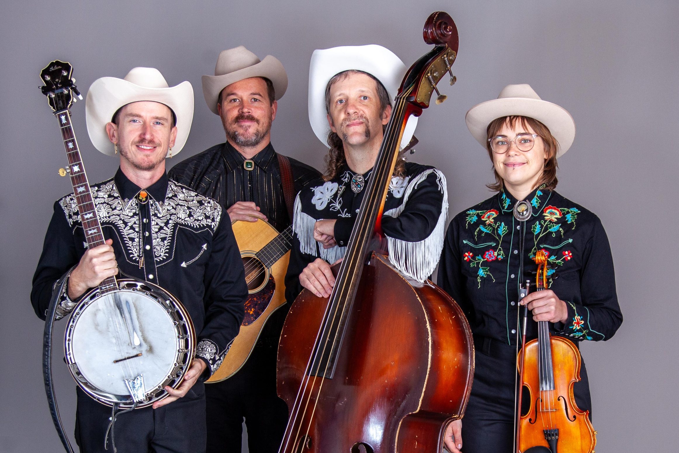 The Jimmy Touzel Bluegrass Explosion – Photo-Rev 2026-01-19 Four musicians in western attire stand with their instruments: a banjo, guitar, upright bass, and violin, against a plain gray background.
