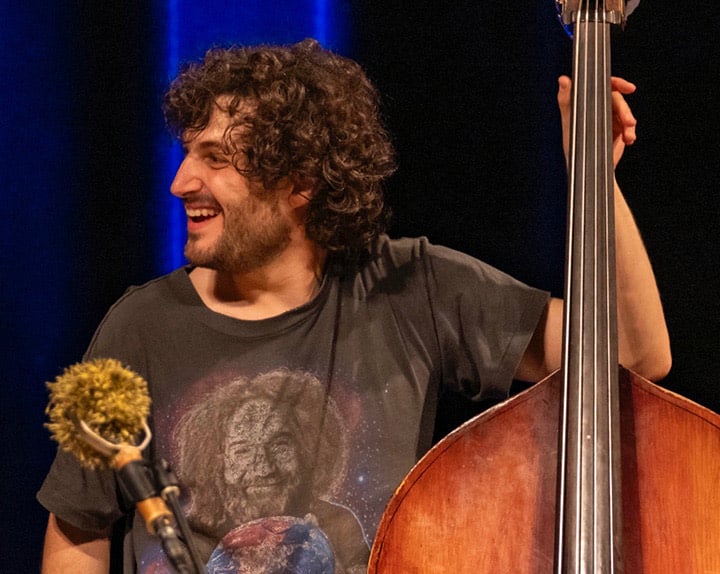 sam-grisman-project_opt A man with curly hair smiles while standing beside an upright bass on a stage, wearing a t-shirt with a graphic design.