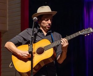 A man wearing a hat plays an acoustic guitar on stage, with a microphone in front of him and purple stage lighting in the background.