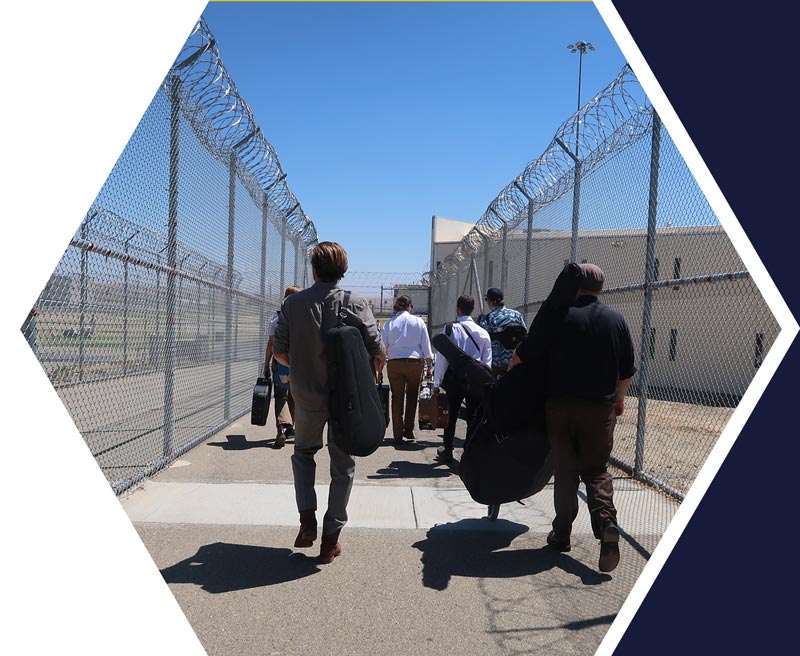 A group of people carrying musical instrument cases walk through a fenced area with barbed wire at an outdoor facility.