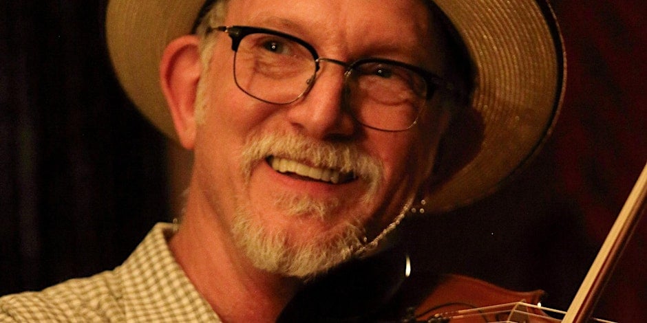 A man with glasses and a straw hat smiles while playing a violin.