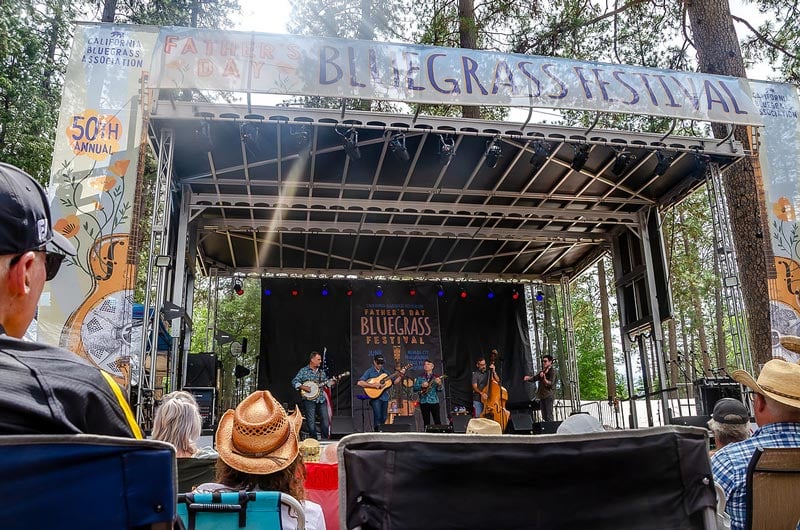 FDF-main-stage-2025 A band performs on stage at the Father's Day Bluegrass Festival, with audience members seated outdoors under trees.