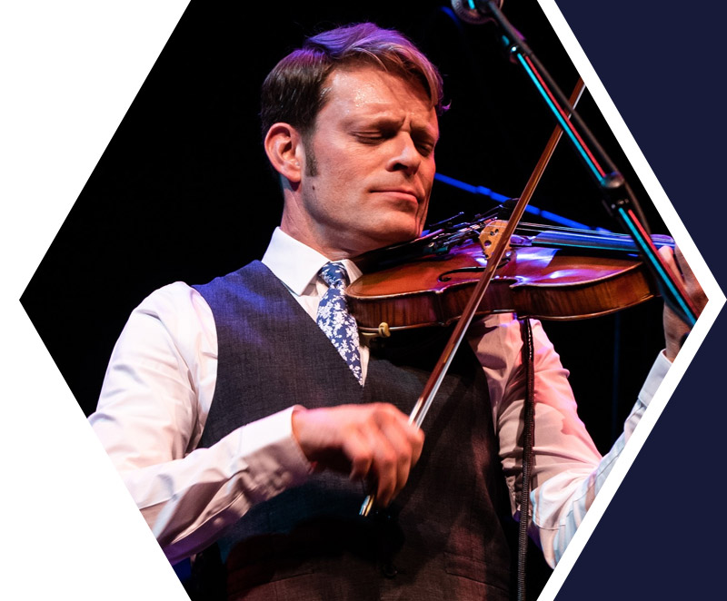 A man in a vest and tie plays the violin on stage, eyes closed, with a microphone in front of him against a dark background.