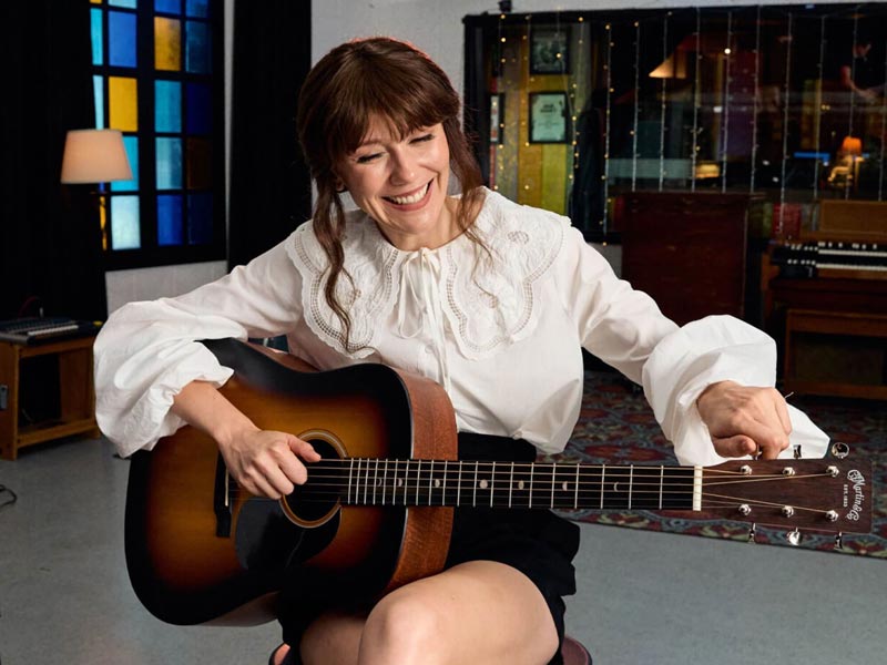 Woman in a white blouse and black shorts tunes an acoustic guitar while sitting indoors, smiling.