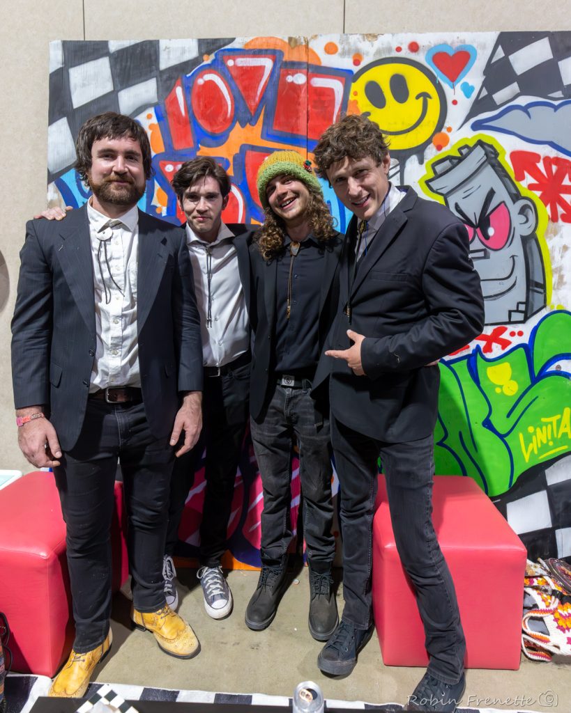 Four men in suits pose together in front of a colorful graffiti wall, with red couches and a can on the floor nearby.
