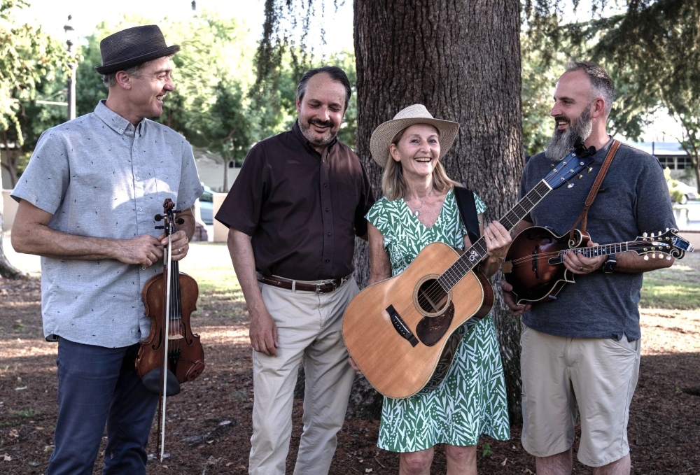 Four adults stand outdoors near a tree, smiling, with musical instruments including a violin, guitar, and mandolin.