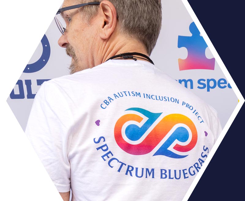 Man wearing a white t-shirt with the logo and text "CBA Autism Inclusion Project Spectrum Bluegrass" printed on the back. Puzzle piece graphic in the background.