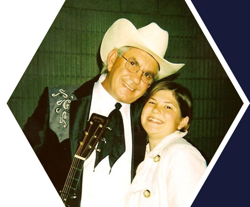 An older man in a cowboy hat and western suit stands smiling with a guitar next to a young woman, both posing for the camera indoors.