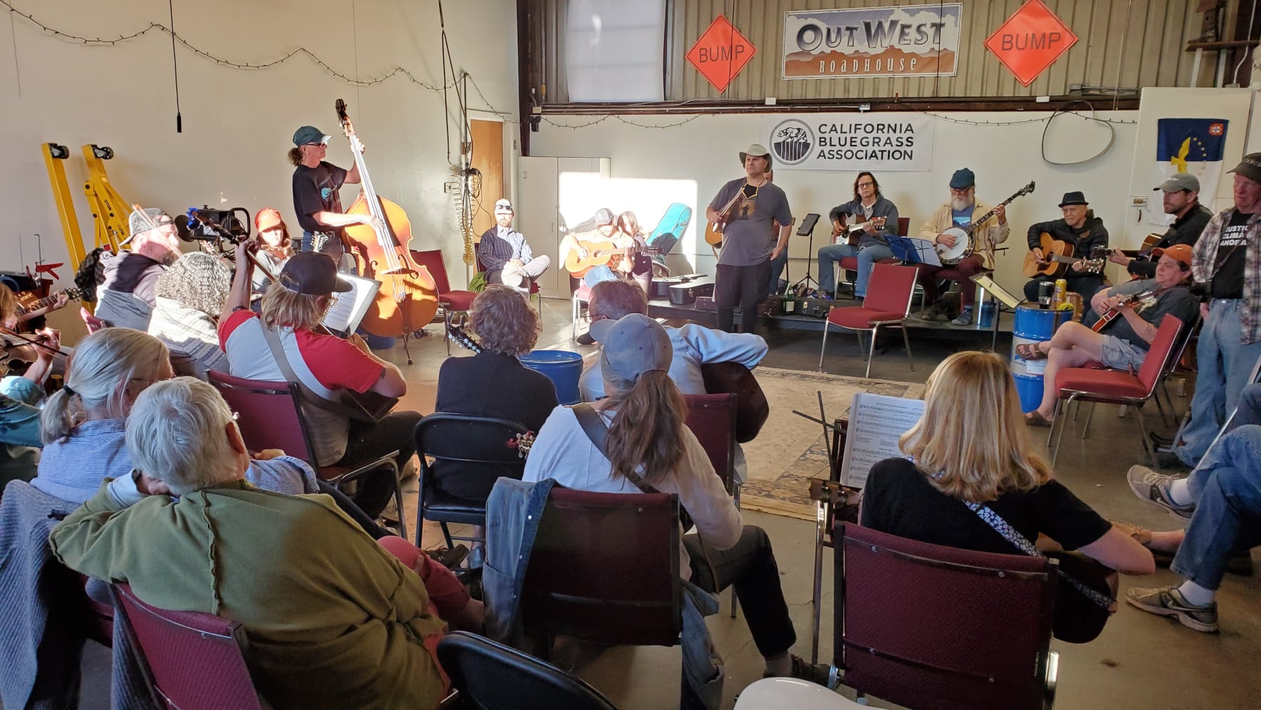 A group of musicians plays string instruments in a semi-circle while an audience watches in a workshop space, with California Bluegrass Association signage in the background.