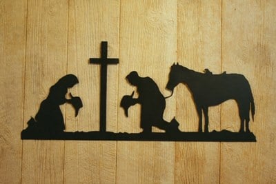 Silhouette of a woman, a man, and a horse with the man and woman kneeling by a cross against a wooden background.