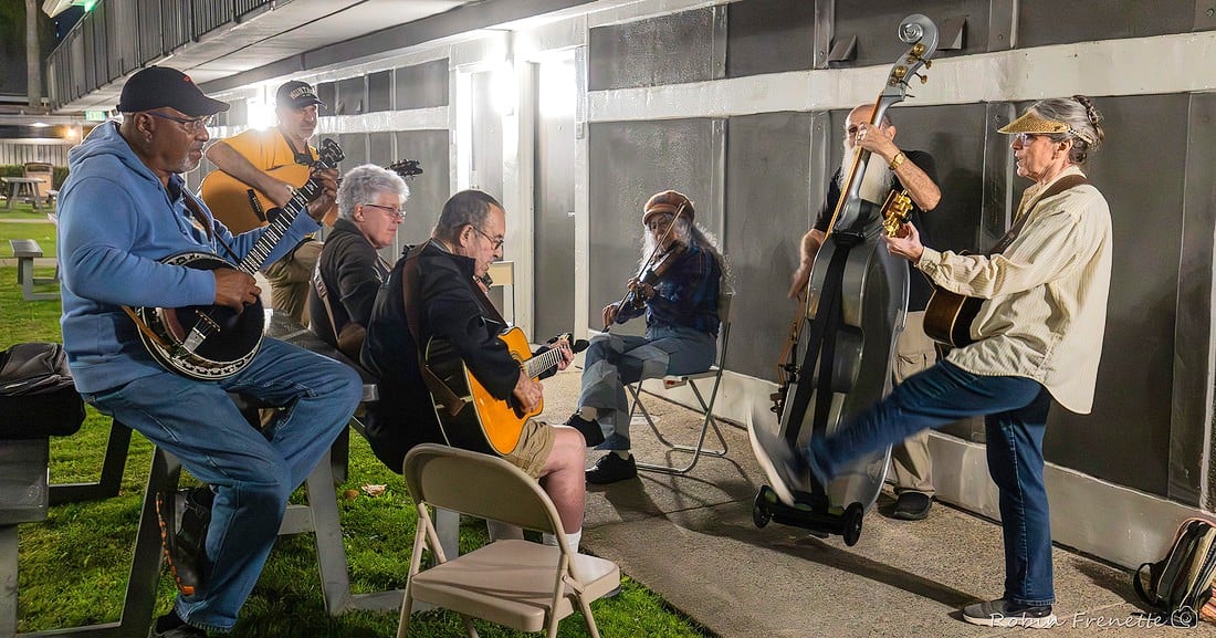 Six people play string instruments and sit in a circle outdoors at night, with one standing musician lifting a leg while playing an upright bass.