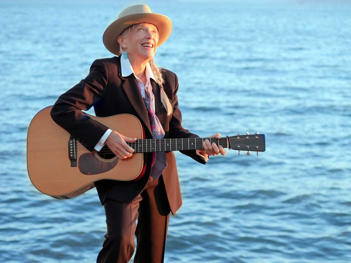 A person wearing a suit, tie, and hat stands by the water, smiling and playing an acoustic guitar.