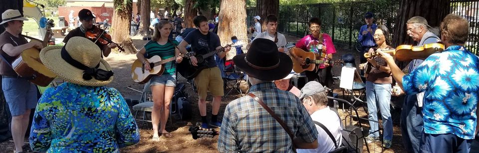 A group of people playing guitars, violins, and other instruments in a circle outdoors under trees at the Santa Clara Valley Fiddlers Association - First Sunday Jam, with some audience members watching.