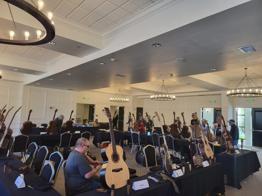 A man sits at a table with papers in a room displaying multiple guitars on stands; several people are walking and observing the instruments.