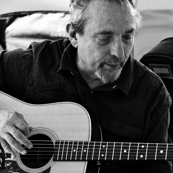 A man with a beard and mustache plays an acoustic guitar, looking down at the instrument. The image is in black and white.