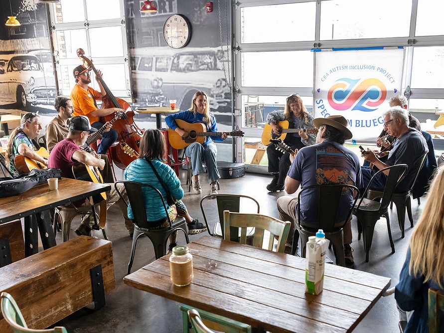 A group of people sit in a circle playing acoustic instruments indoors, with a "Spectrum Bluegrass" banner visible in the background.