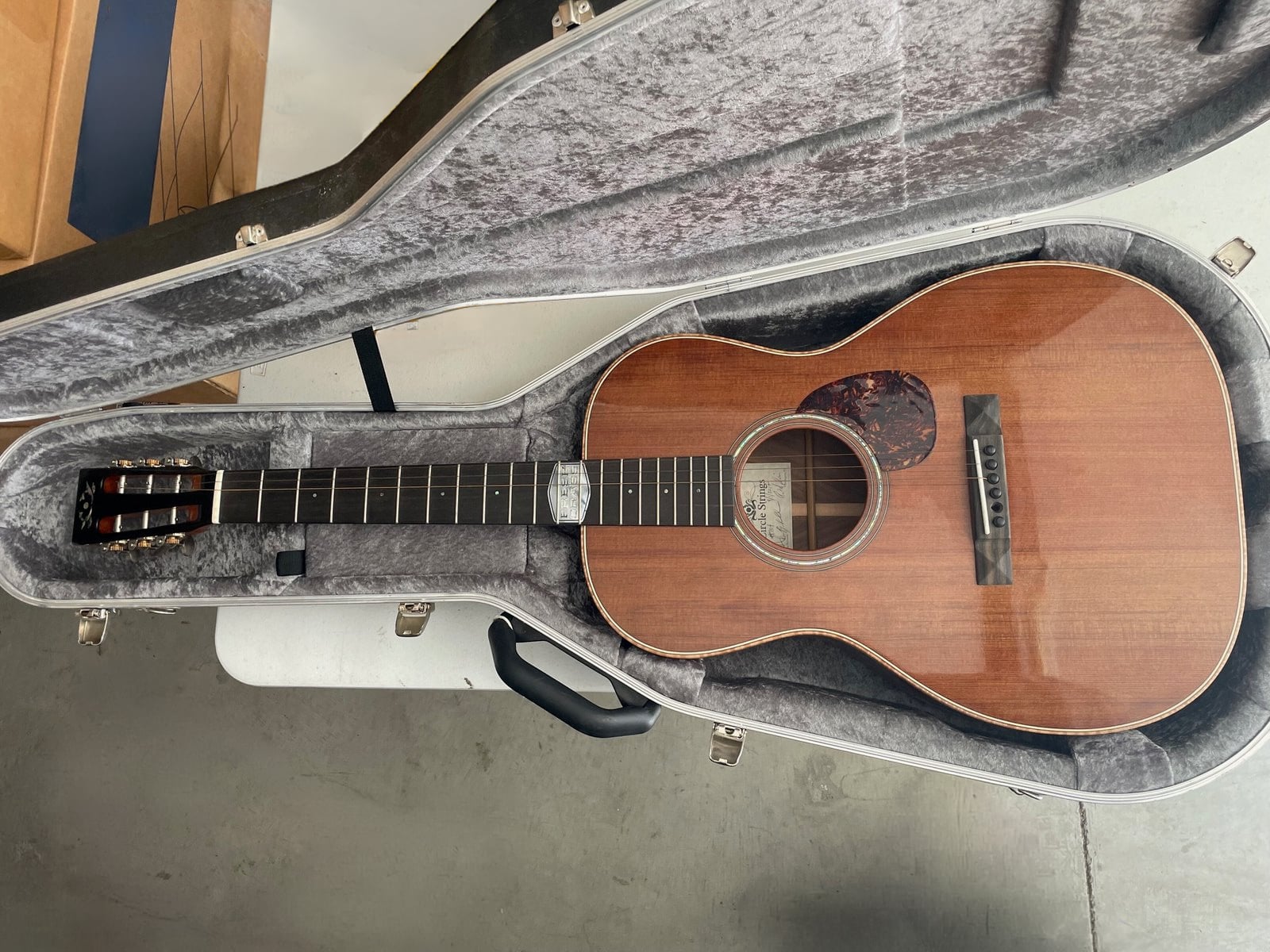 A wooden acoustic guitar with nylon strings is placed inside a grey, plush-lined hard case on a concrete floor.