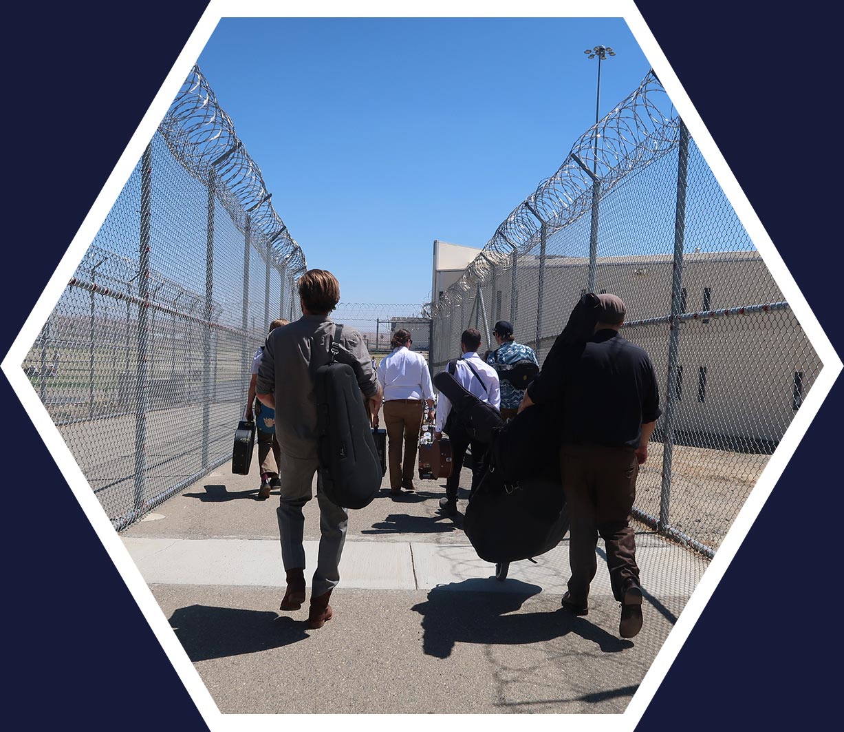 A group of people carrying musical instrument cases walk through a fenced, barbed-wire corridor at a secure facility under a clear blue sky.