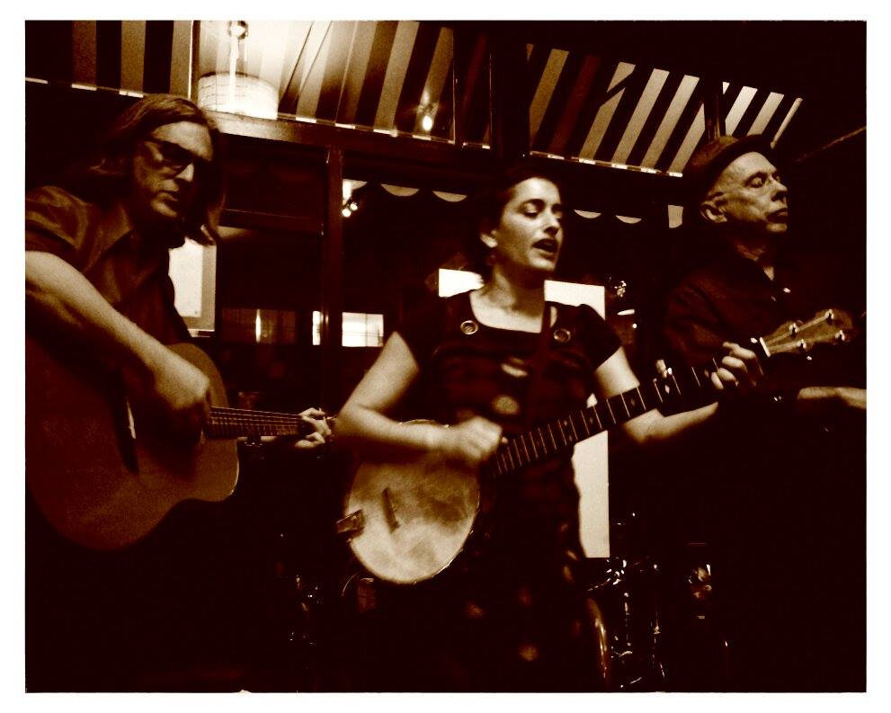 Three musicians perform indoors; one plays guitar, one sings and plays banjo, and one plays another string instrument. The photo has a sepia tone.