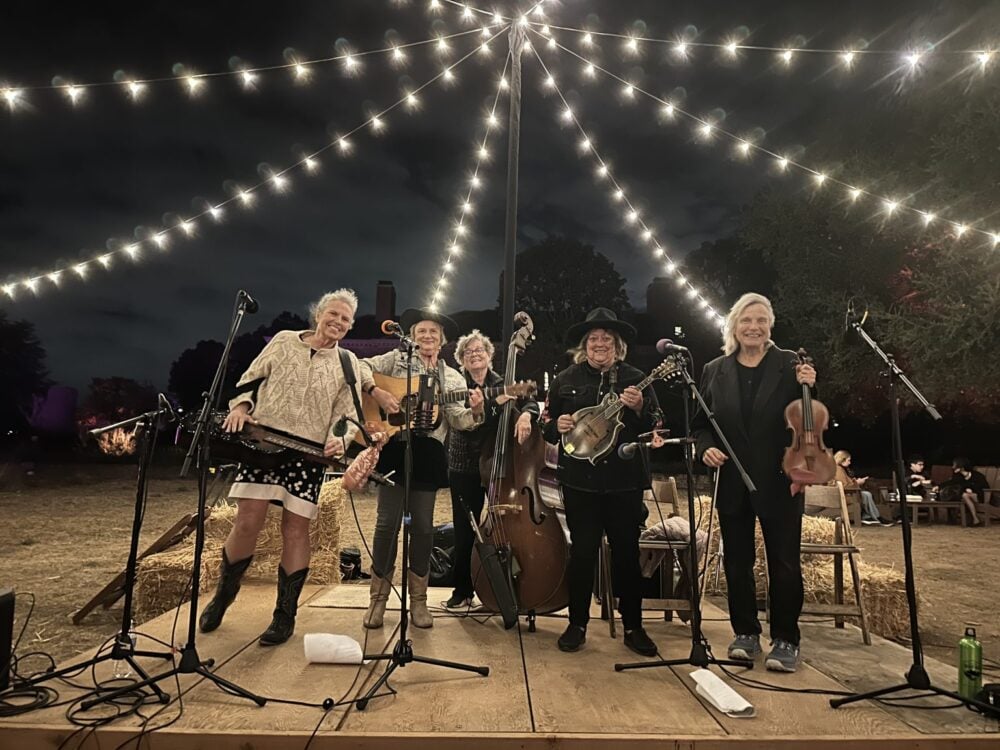 Five musicians with string instruments stand smiling on an outdoor stage under string lights at night, with hay bales and trees in the background.