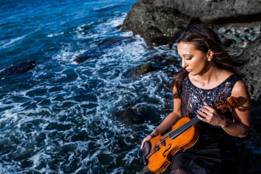 KATIE GLUMAC A woman in a black dress holds a violin while sitting on rocks by the ocean, with waves crashing behind her.