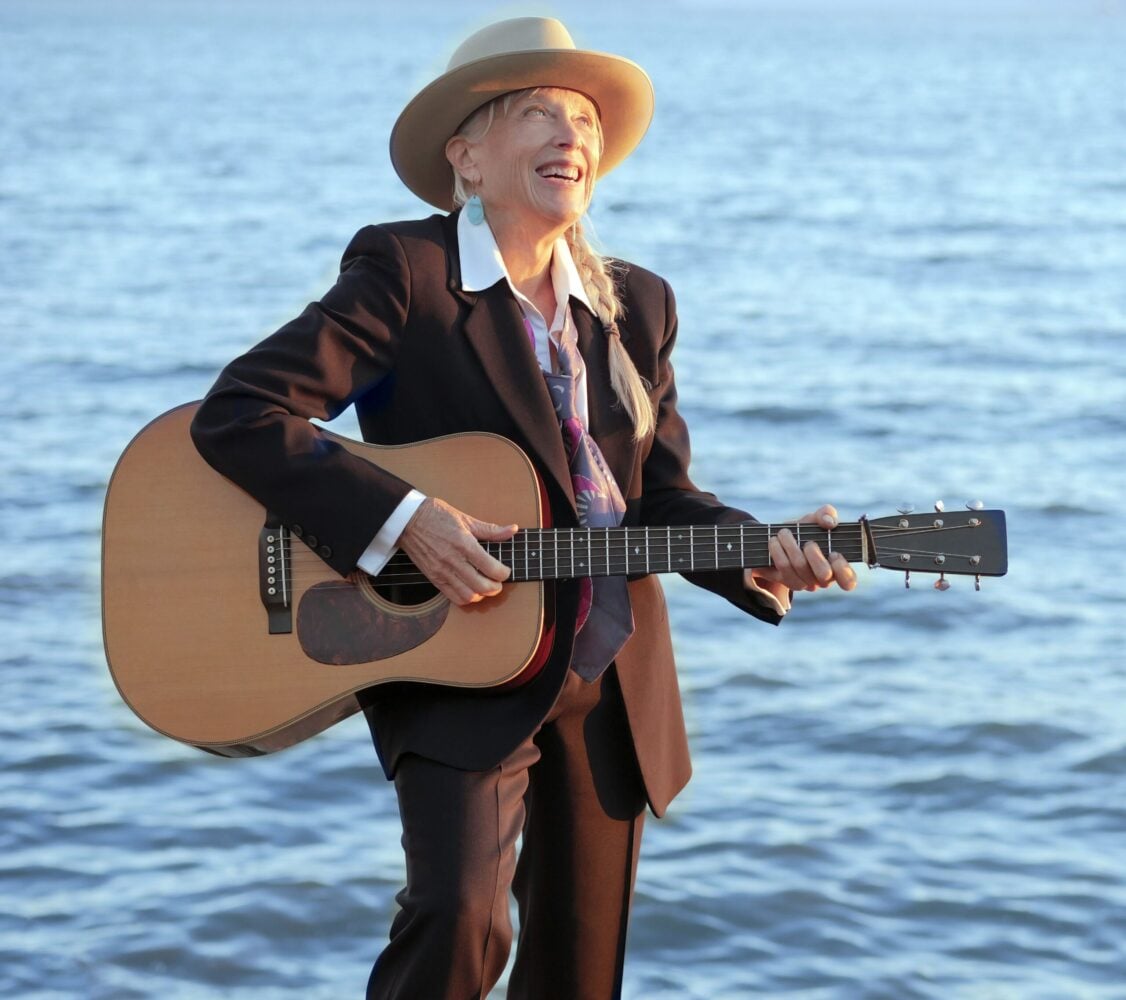 A person in a suit and hat stands by the water, smiling and playing an acoustic guitar.