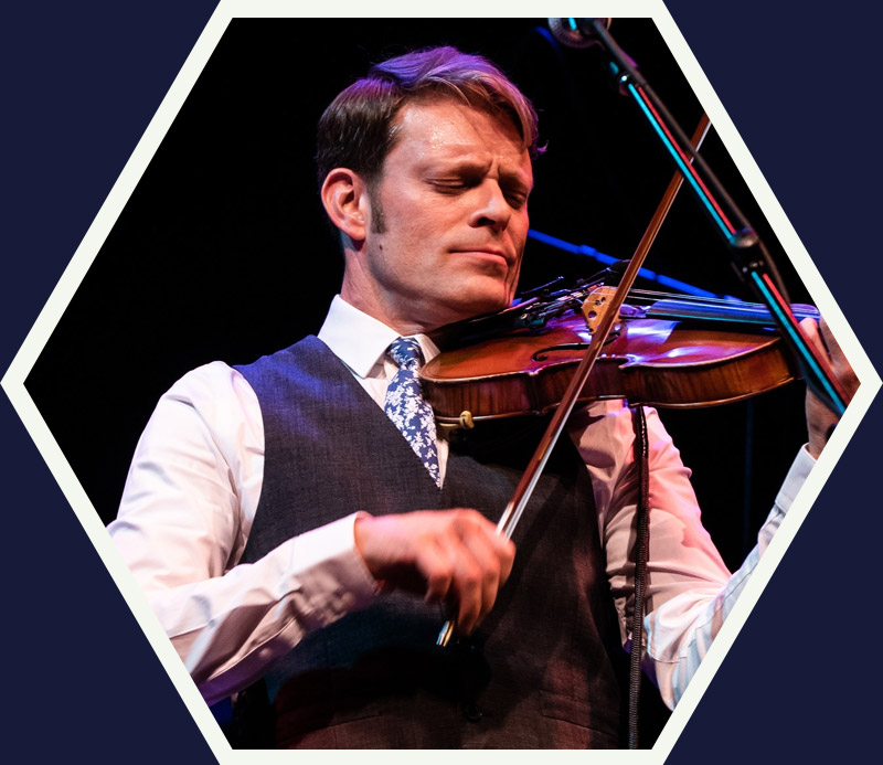 A man in a vest and tie plays a violin on stage with a microphone nearby, framed by a hexagonal border.