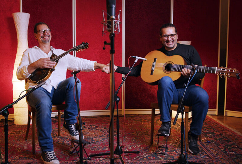Two men sit in a recording studio holding musical instruments, smiling and shaking hands, with microphones in front of them.