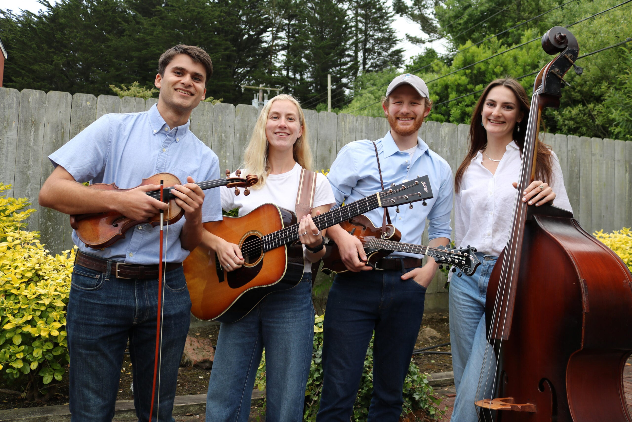 Four people stand outdoors holding musical instruments: a violin, guitar, mandolin, and upright bass, with greenery and a wooden fence in the background.