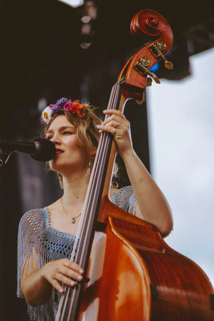 A woman wearing a flower crown plays a double bass and sings into a microphone on an outdoor stage.