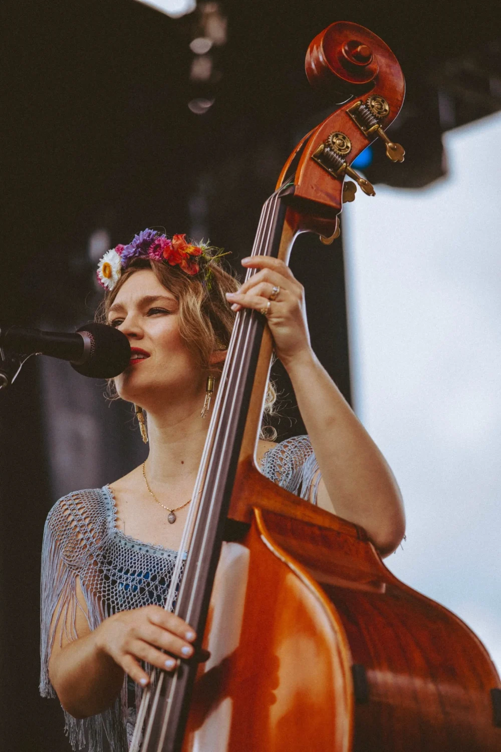 A woman wearing a flower crown plays a double bass and sings into a microphone on an outdoor stage.