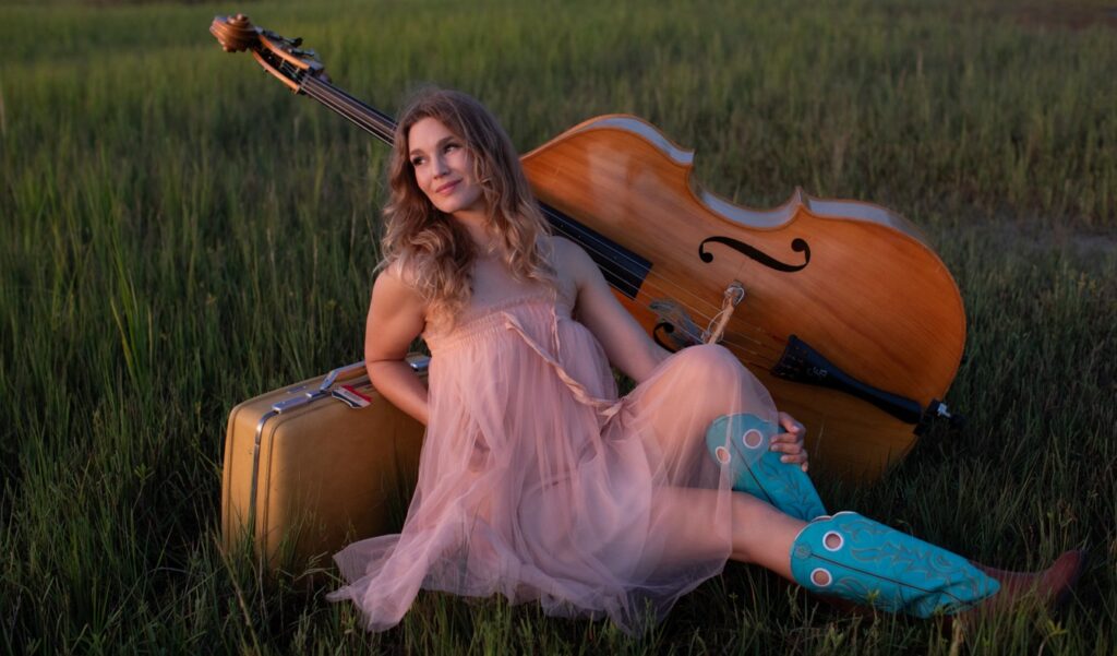 Woman in a sheer pink dress and turquoise boots sits on grass, leaning against a suitcase and an upright bass at dusk—capturing the spirit of Shelby Means Band @ Otter Opry in an outdoor field.