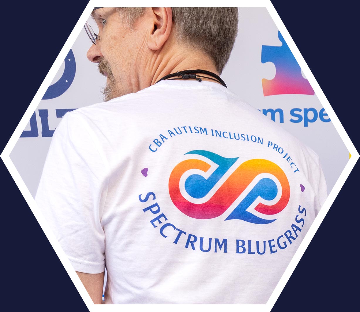 A man faces away, wearing a white t-shirt with the "CBA Autism Inclusion Project Spectrum Bluegrass" logo in blue, orange, and pink on the back.