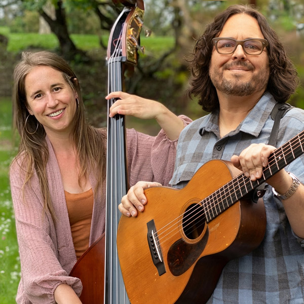 Two people standing outdoors, one holding an upright bass and the other holding an acoustic guitar, both smiling at the camera.