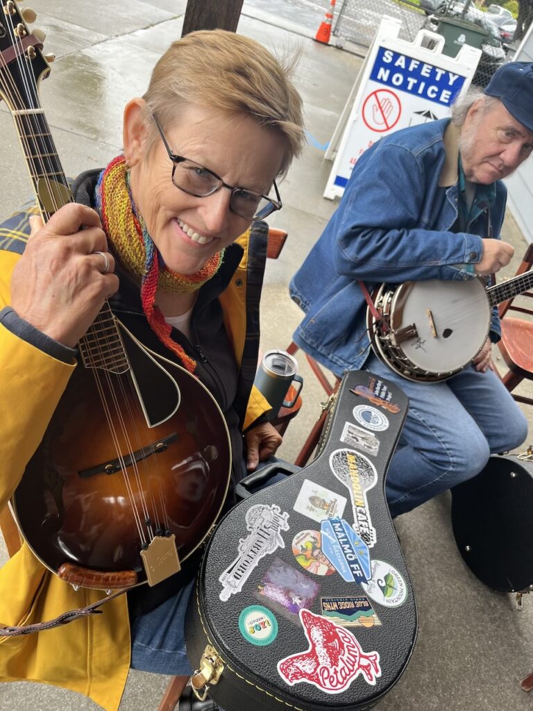 Two people sit outdoors with string instruments; one holds a mandolin and smiles at the camera, while the other plays a banjo. A sticker-covered case and safety sign are visible.