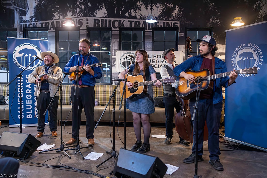Five musicians perform on stage with string instruments at a California Bluegrass Association event, standing in front of banners and microphones inside an indoor venue.
